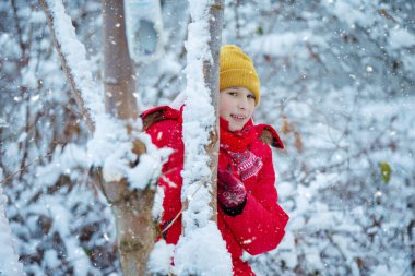 Kışın giyinmiş bir çocuk karlı bir ağaç gövdesinin arkasından dışarı bakar. Kar yağıyor..