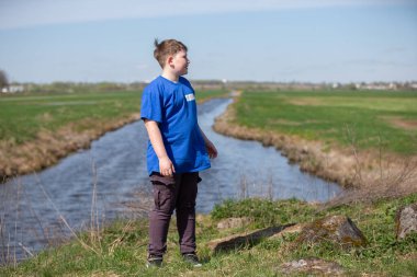 Genç bir çocuk bir nehrin ve yeşil bir çayırın arkasında duruyor..