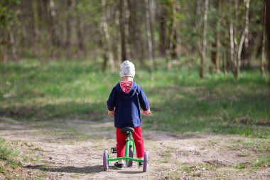 Küçük bir çocuk orman yolunda üç tekerlekli bisiklet sürüyor..