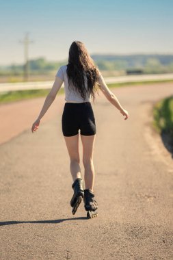 Yolda bir kadın paten kayıyor. Beyaz gömlek ve siyah şort giyiyor..