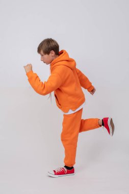 Turuncu tulumlu ve kırmızı ayakkabılı genç bir çocuk. Kollarını açmış koşuyormuş gibi yapıyor..
