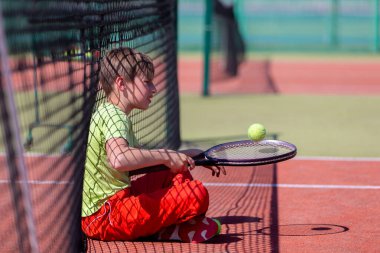 Bir çocuk ağda oturur ve tenis raketiyle topa vurur..