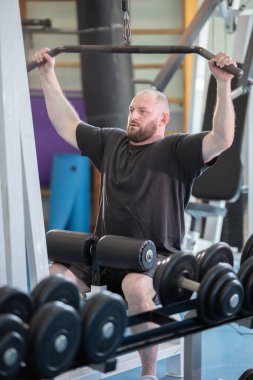 Bir adam spor salonunda ağırlık kaldırıyor. O, siyah bir gömlek ve şort giyiyor. Spor salonunda bir sürü ekipman var. Buna göğüs presi ve halterler de dahil.