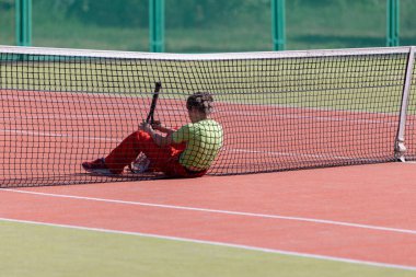 Çocuk elinde raketle tenis ağının başında oturuyor. Yarışmayı kaybetti ya da antrenmandan yoruldu..
