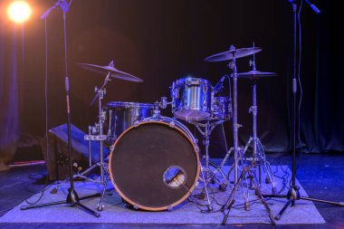 Sahnede mavi sahne ışıklarıyla aydınlatılmış bir mikrofon ve davul seti var..