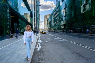 Şık bir kadın, yansıtıcı binalarla dolu hareketli bir şehir caddesinde yürüyor. Güneş ışıl ışıl parlıyor, günlük kıyafetini aydınlatıyor ve çevresindeki hayat dolu şehir hayatını aydınlatıyor..