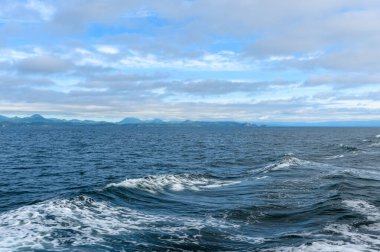 Yumuşak dalgalar okyanus boyunca dalgalanmalar yaratırken, bulutlar yukarıda sürükleniyor. Uzak dağlar ufukta yükseliyor, sakin bir kıyı manzarasını yansıtıyor..