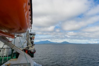 Ufukta yemyeşil adalar belirirken sakin suların üzerinde bir gemi süzülüyor. Yumuşak bulutlar, açık denizde barışçıl bir deniz manzarası yaratıyor..