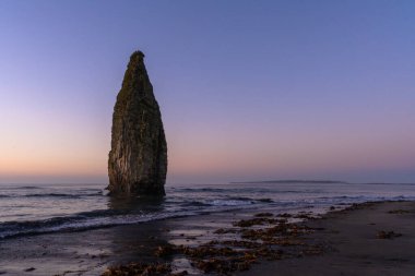Sakin bir Rus plajına alacakaranlık çökünce sakin denizden çarpıcı bir kaya oluşumu etkileyici bir şekilde yükselir. Yumuşak dalgalar, deniz bitkileriyle bezenmiş kumlu kıyı şeridine nazikçe çarpar..