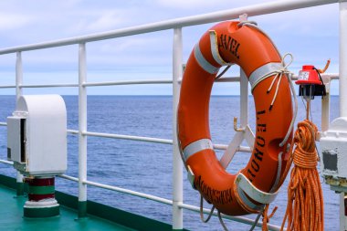 Canlı bir turuncu can simidi, sakin suya tepeden bakan bir geminin korkuluklarına sabitlenir. Gökyüzü parçalı bulutlu, Rusya 'da sakin bir deniz atmosferi sergiliyor..