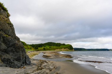 Sakin sular uçurumlarla kaplı engebeli bir kıyı şeridiyle buluşur, uzaktaki yeşil tepeler yükselir ve bulutlar yumuşak, bulutlu bir gökyüzü yaratır. Doğa barışçıl bir ortamda ortaya çıkar..