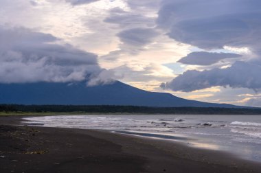 Kıyı şeridi boyunca uzanan kara kumlar dalgalar yavaşça kıyıya çarpıyor. Büyük bir dağ, alacakaranlıkta karamsar bir gökyüzünün altında bulutlarla kaplanmış arka plana hükmediyor..
