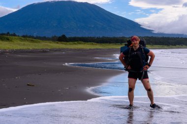Bir gezgin, doğanın güzelliğiyle çevrili ıslak bir kumsalda duruyor. Arka planda görkemli bir dağ yükselir ve gökyüzü dramatik bulutlarla doludur..