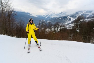 Parlak sarı kayak takımlı bir kadın, karlı dağ zirvelerine karşı poz veriyor. Kış tatili atmosferi ve aktif turizm.