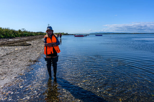 A person stands in shallow water wearing an orange safety vest, enjoying the sunny day along the calm shore. Colorful boats dot the picturesque landscape in the distance.