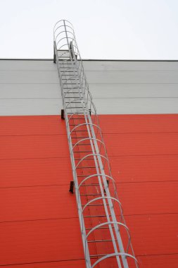 Uzun metal bir merdiven, bulutlu bir gökyüzüne çıkan, çarpan turuncu bir duvara doğru uzanır. Yükseliş ve olasılığın sembolü olarak duruyor..