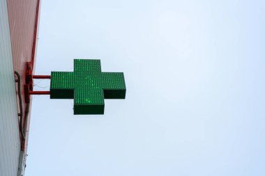 Bulutlu gökyüzünü aydınlatan yeşil bir haç işareti, bir eczane binasında belirgin bir şekilde sergileniyor, sağlık hizmetlerini ve toplumdaki desteği simgeliyor..