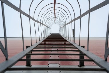 Bir insan, bulutlu bir günde, parlak, açık gökyüzüne uzanan kırmızı tuğladan bir duvara doğru uzun metal bir merdiven tırmanır..