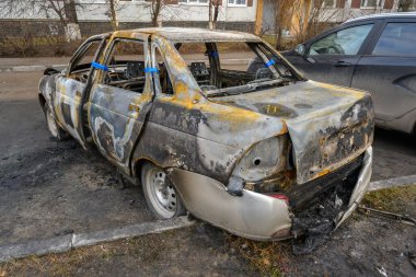 Yanmış ve hasarlı bir araba bir şehir caddesinde terk edilmiş durumda. Yanmış iskeleti yangının sonrasını gösteriyor. Yakınlardaki araçlara dokunulmamış..