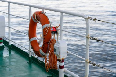 Canlı bir turuncu yaşam halkası, bir teknenin korkuluklarından sarkan, denizdeki güvenlik önlemlerini yansıtan. Sakin sular sakin bir zemin oluşturur. Huzurlu bir yolculuk olacağını ima eder..