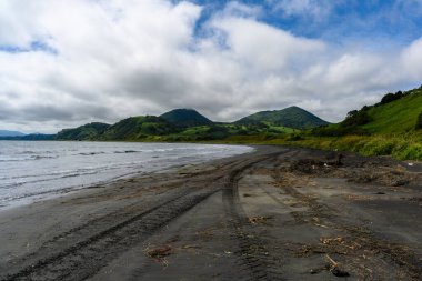 Sakin bir sahil sahnesi, yumuşak dalgaların karanlık kumu yalayıp yutmasıyla ortaya çıkar. yemyeşil tepeler arkaplanda görkemli bir şekilde yükselir, parçalı bulutlu bir gökyüzünün altında.