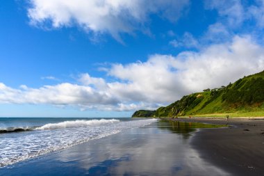 Dalgalar yavaşça kıyıya vururken bulutlar canlı yeşil bir tepenin üzerinde yüzüyor. İnsanlar ıslak kumda gezinir, okyanus kenarında huzurlu bir günün tadını çıkarırlar..