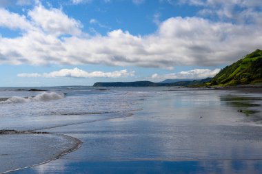 Parlak güneş ışığı, yumuşak dalgaların kıyı şeridini öptüğü, tüylü bulutlarla dolu güzel bir gökyüzünün altındaki bereketli tepelerin çerçevelediği sakin sahili aydınlatır..