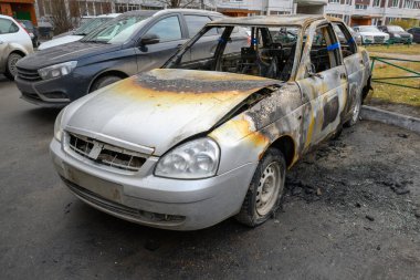 Kömürleşmiş bir araba şehir caddesinde terk edilmiş duruyor. Vücudu yanmış ve paslanmış, diğer park etmiş araçlarla çevrili. Bölge sessiz ve unutulmuş görünüyor..