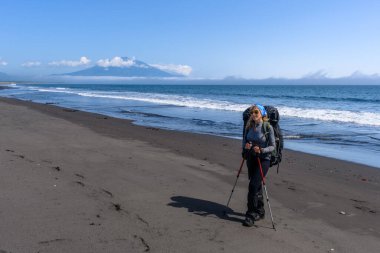 Tutkulu bir yürüyüşçü, arka planda yükselen bir dağ ve açık bir günde sahile vuran dingin okyanus dalgalarıyla Rusya 'nın kara kumlu plajında yürüyor..