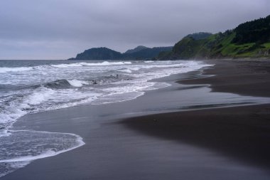 Rusya 'da sakin bir plajda koyu kumlar ve dalgalar bulunur. Manzara bereketli tepeler tarafından çerçevelenmiş, doğa denizle buluşurken sakin bir atmosfer yaratıyor..