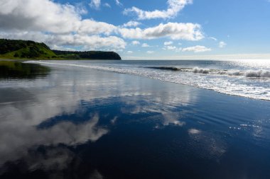 Dalgalar, parlak mavi bir gökyüzünün altında sakin, siyah bir kumsala nazikçe çarpar. Pofuduk bulutlar suyu yansıtıyor ve bu pitoresk Rus manzarasında huzurlu bir atmosfer yaratıyor..