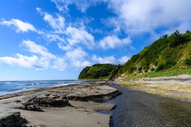 Sahilde uzanan kumlu güzel bir sahil var. Arkaplanda yemyeşil kayalıklar yükseliyor. Mavi gökyüzü kabarık beyaz bulutlarla dolu..