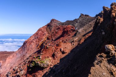 Rusya 'daki engebeli bir volkanik dağın tepesinde, canlı kırmızı ve koyu kaya oluşumları açık mavi gökyüzüne karşı yükseliyor, doğanın elementlerinin ve dokularının çarpıcı kontrastını gözler önüne seriyor..