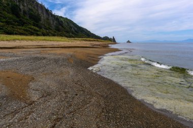 Sahil boyunca uzanan sakin bir sahil kıyıya doğru yumuşak dalgalar dalgaları ile birlikte. Yeşillik ve kayalık çıkıntılar dingin sulara karşı çarpıcı bir zemin oluşturur..