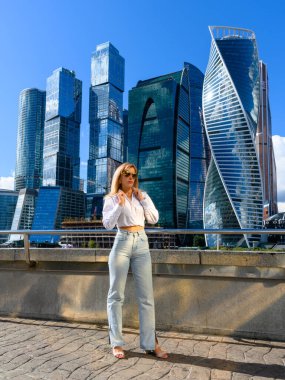 Şık bir genç kadın, şehrin canlı mimarisini gözler önüne seren gökdelenlerin önünde duruyor. Bu güzel günde güneş parlıyor..