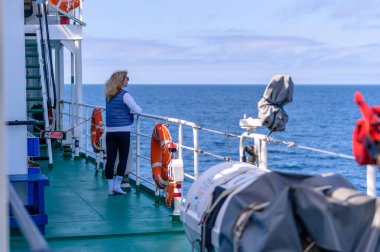 Bir gezgin, bir geminin güvertesinde durmuş, uçsuz bucaksız mavi okyanusa bakıyor. Yaşam şamandıraları, açık mavi bir gökyüzünün altında hafif dalgaların tekneye çarpmasıyla yan yana dizilirler..