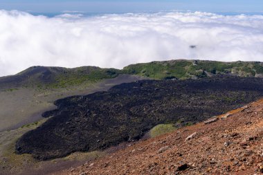 Çarpıcı bir siyah lav tarlası engebeli araziye yayılır, parlak mavi bir gökyüzünün altında yemyeşil yemyeşil ile çevrilidir. Bulutlar volkanik manzaranın üzerinde sakin bir şekilde süzülür..