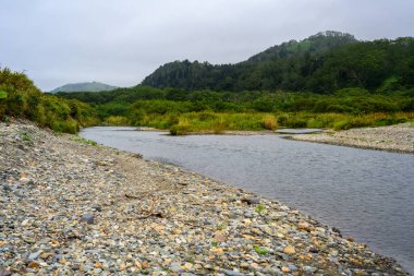 Huzurlu bir nehir, tepelerle çevrili canlı yeşil bir vadiden yavaşça akar. Yumuşak bulutlar, alacakaranlıkta huzurlu bir atmosfer yaratır..