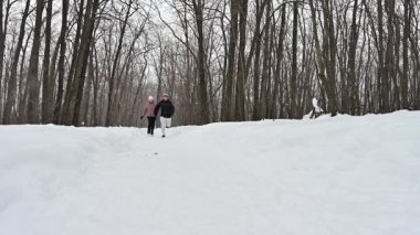 Neşeli bir çift, karlı bir patika boyunca el ele koşar, uzun ağaçlarla çevrili, hafif bir kar altında. Kış aktivitelerinin neşesini kucaklarken kahkahaları havayı dolduruyor..