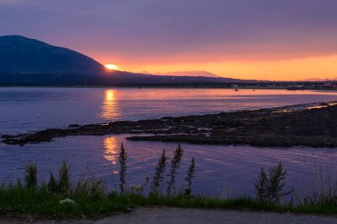 Güneş sakin suların üzerinde batarken, turuncu ve mor yumuşak renkler gökyüzünü doldurur. Uzak dağların gölgeleri sakin bir akşam sahnesi için sakin bir zemin oluşturur..
