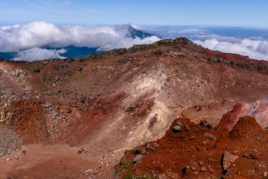 Kayalık volkanik arazinin nefes kesen manzarası yerden yükselen canlı renkler ve buhar içerir. Yoğun bulutlar havada süzülürken, uzak tepeler çarpıcı bir zemin oluşturur.