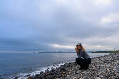 Kayalık bir sahilde çömelmiş, yaklaşan yağmuru işaret eden gri gökyüzünün altındaki sakin denizi dikkatle seyreden bir kadın. Huzurlu manzara yansımayı ve barışı davet eder..