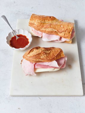 Öğle yemeğinde baget, mozzarella, marul ve jambondan yapılmış taze sandviçler sergileniyor. Yakınlarda bir tabak domates sosu duruyor..