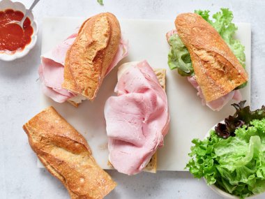 Taze malzemelerle yapılan sağlıklı sandviçler bir yemek masasına konur. Ekmek, marul, mozzarella ve jambon hazır.