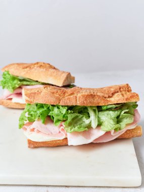 Ekmek, mozarella peyniri, marul ve jambonlu iki sandviç beyaz bir yüzeye oturur..