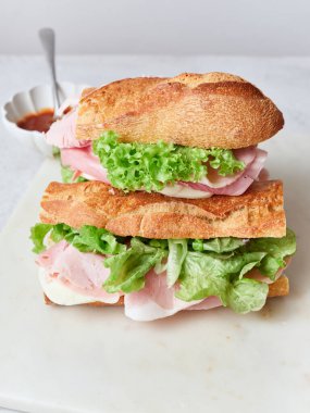 Ekmek, mozarella peyniri, marul ve jambondan yapılmış sağlıklı bir sandviç. Taze dilimlenmiş portakal ve çırpılmış yumurta yakında. Bir kase domates sosu da var..