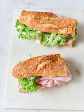 İki dilimlenmiş sandviç, beyaz bir yüzeye salata, jambon ve mozzarella peyniri ile konur..