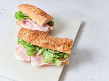 İki sandviç mozzarella peyniri, jambon ve marulla dolu beyaz bir tabakta oturur. Yakınlardaki bir masada portakallar ve çırpılmış yumurtalar var. Bu sahne basit bir yemek düzenini yakalar..