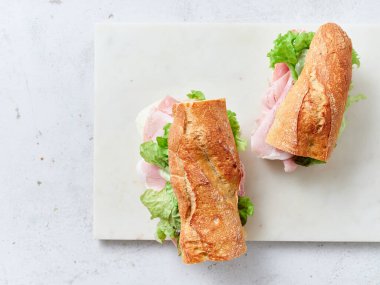 Öğle yemeği veya kahvaltı masasında baget, mozzarella peyniri, jambon ve maruldan yapılmış dilimlenmiş sandviçler var. Taze portakallar ve çırpılmış yumurtalar da masada..