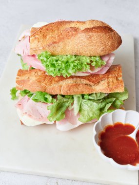 Bir öğle yemeği veya kahvaltı masasında jambon, mozzarella peyniri ve marul dilimleri ve çırpılmış yumurtalarla dolu sağlıklı bir baget sandviç sergilenir..